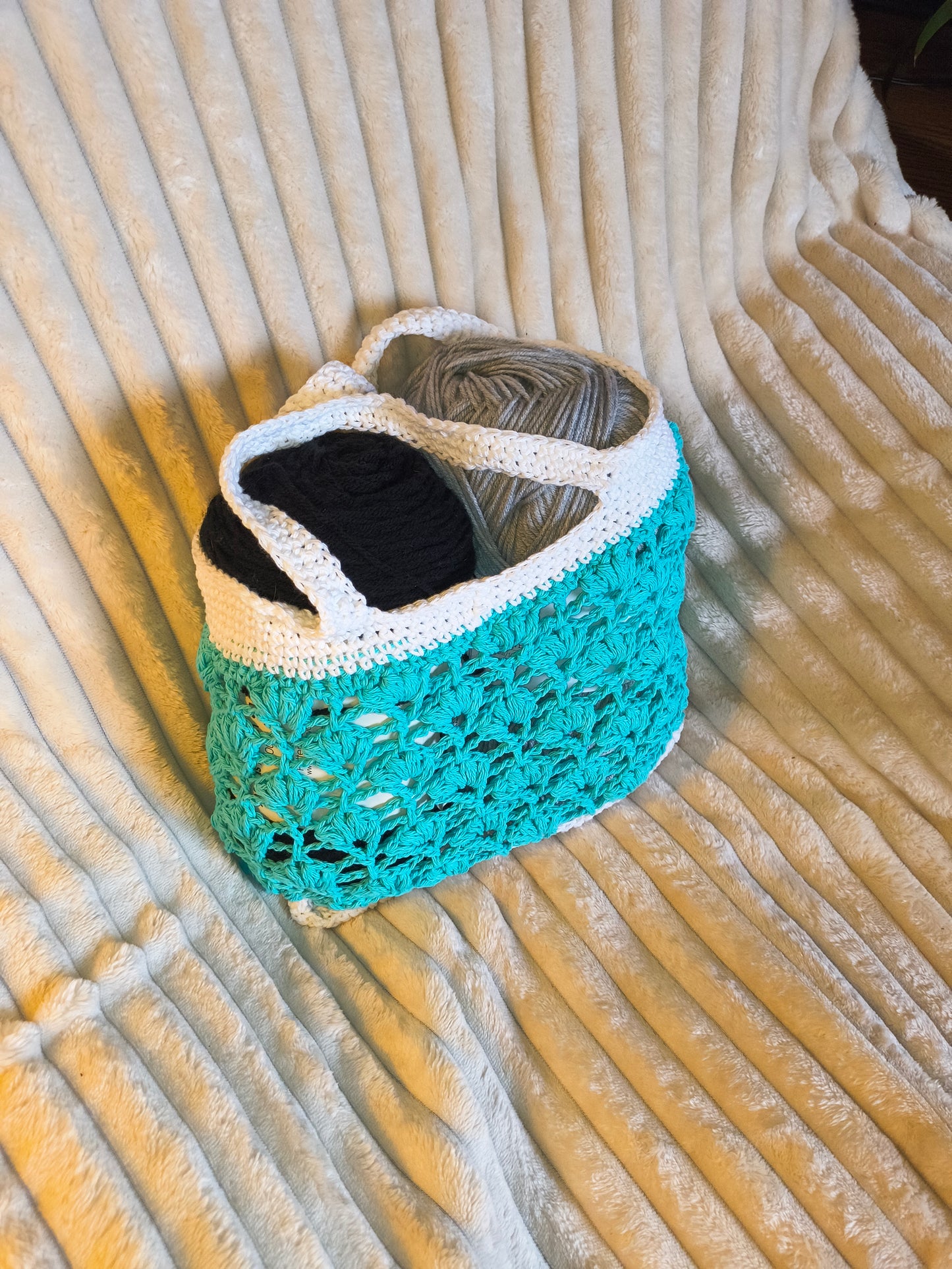Handmade teal and white crochet market bag made from 100% cotton with lace stitch design and soft handles, displayed on a cream textured blanket