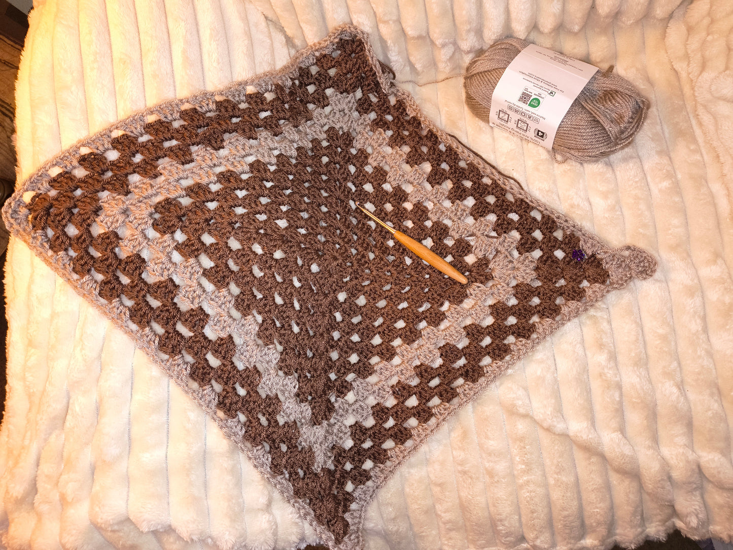 Crochet baby blanket in progress using brown and neutral yarn, featuring an open granny square pattern with a crochet hook and yarn skein resting on a soft cream blanket.
