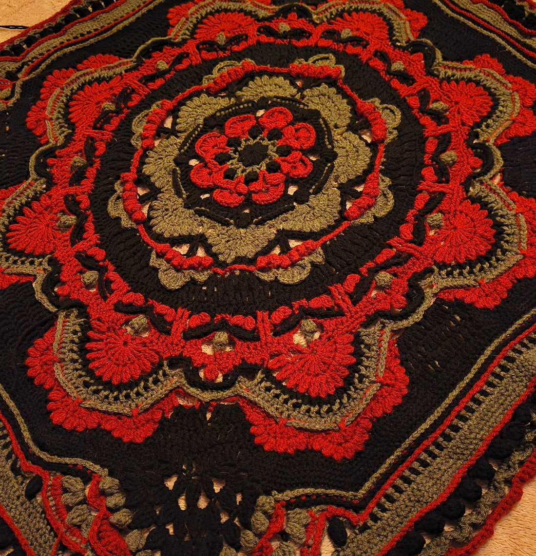 Close-up of a handmade crochet mandala blanket in progress featuring bold red, black, and gray tones with intricate textured stitches and layered floral-inspired design.