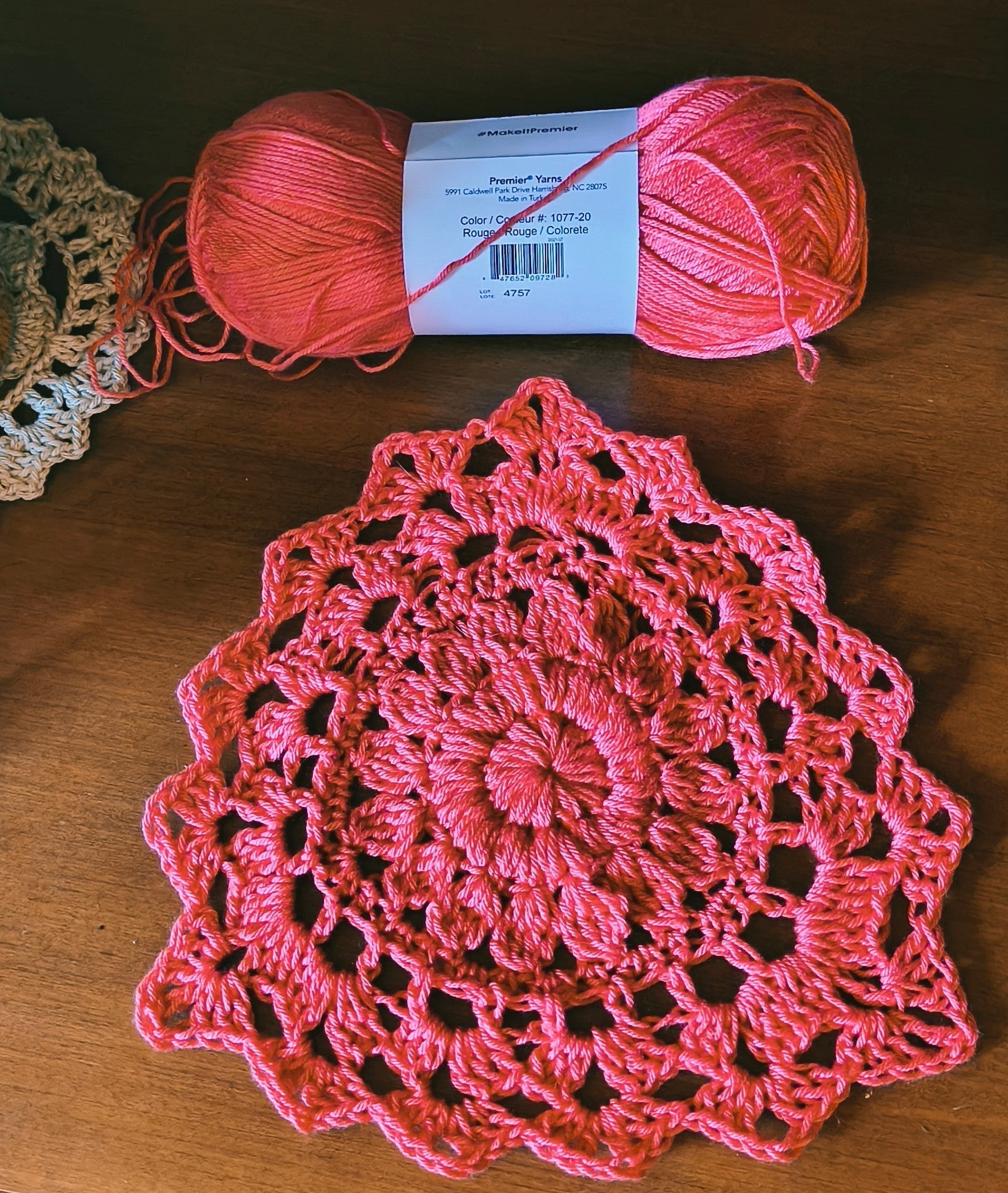 Close-up of a handmade mandala-inspired crochet cup in bright rouge pink yarn resting beside a skein of Premier Yarns Bamboo Fair. The design features textured puff stitches, lace details, and layered floral petal shaping on a wooden surface.