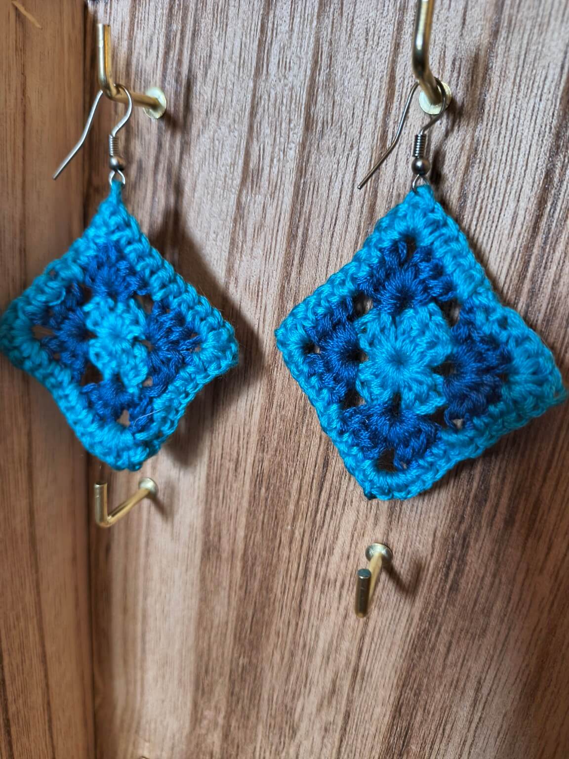 Handmade turquoise blue crochet granny square earrings in a diamond shape with silver-tone hooks, displayed against rustic wood.