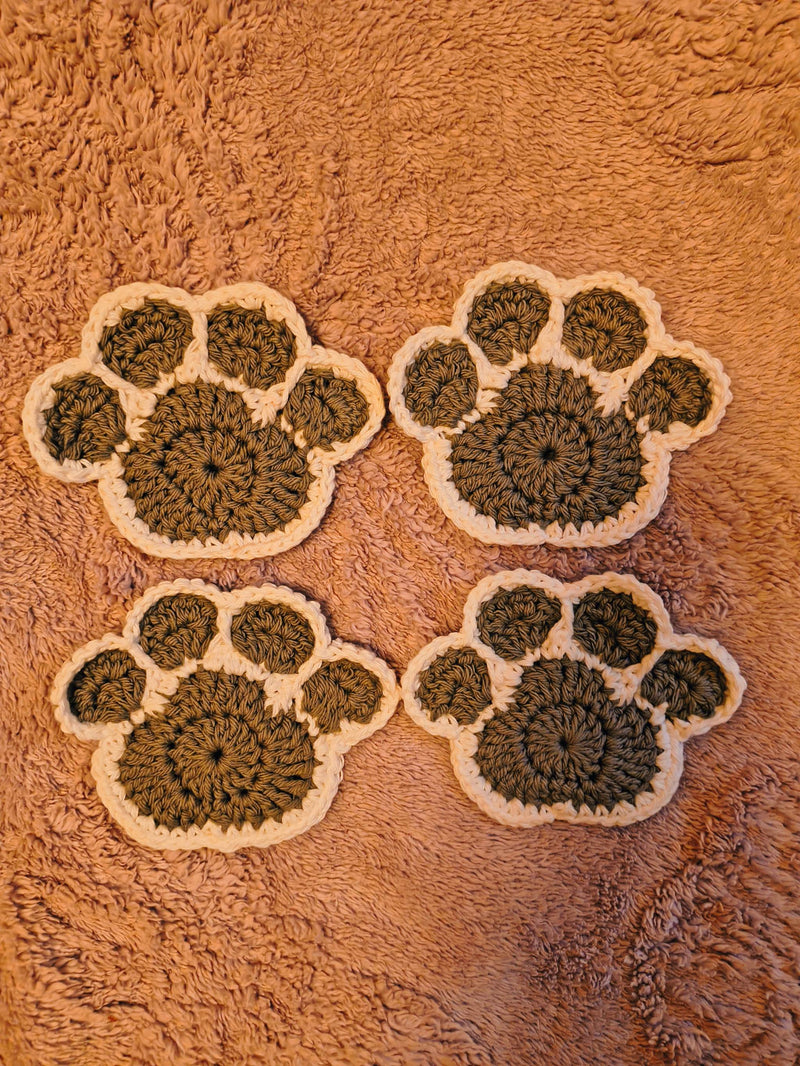 Four crocheted paw print coasters with gray centers and white borders, arranged on a tan background.