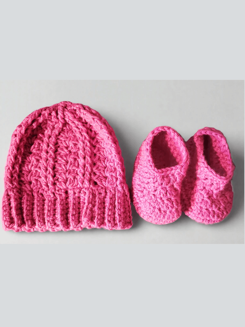Pink crocheted baby hat with a ribbed brim beside a matching pair of baby booties on a light background.