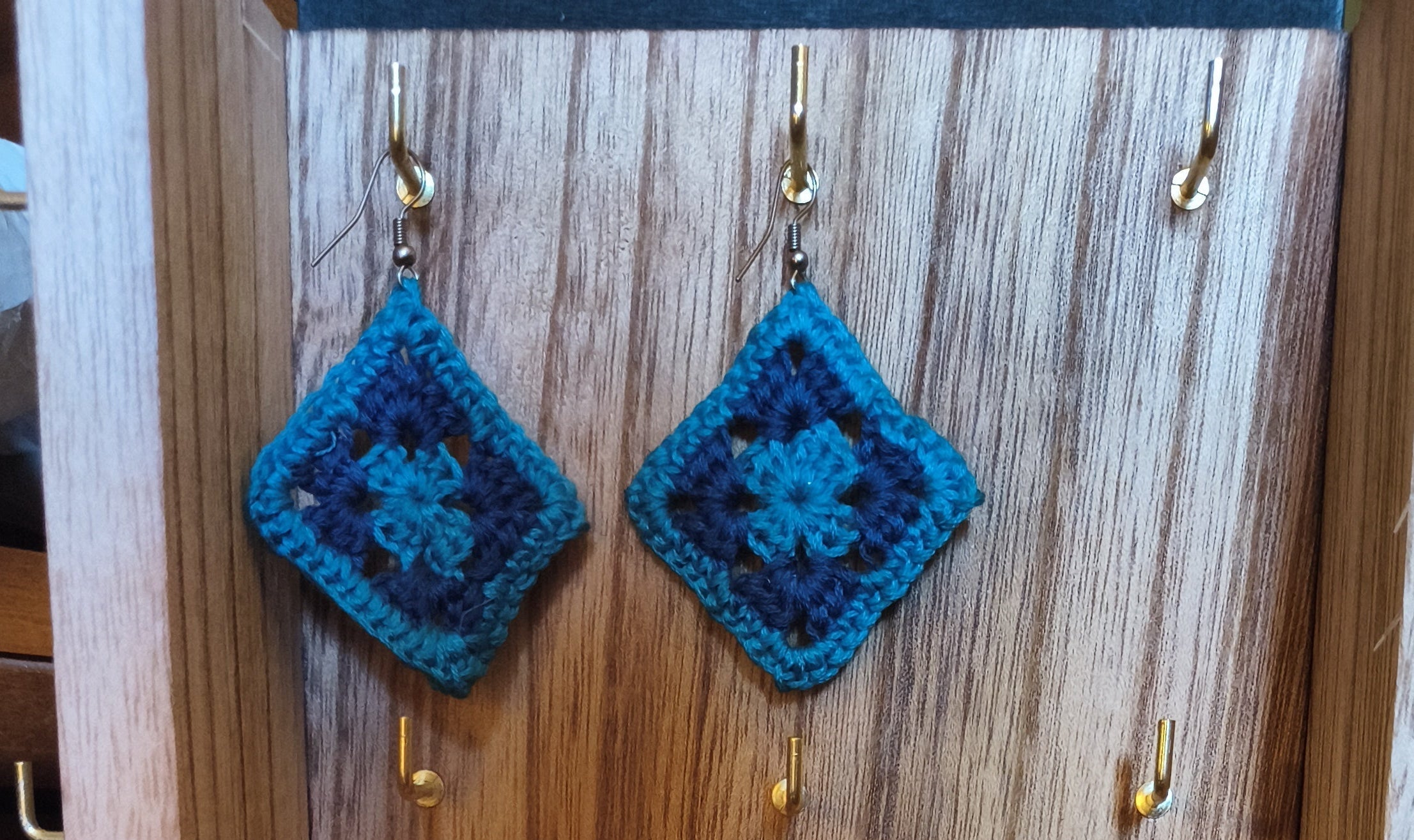 Handmade crochet granny square earrings in teal and blue tones displayed on a wooden jewelry stand.