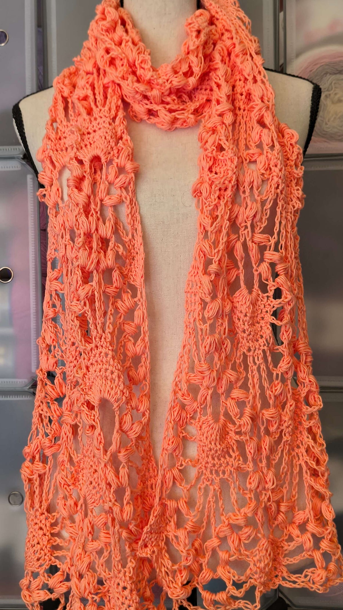 A bright coral orange crochet cowl scarf draped over a dress form mannequin, with a blurred background of storage bins containing yarn.