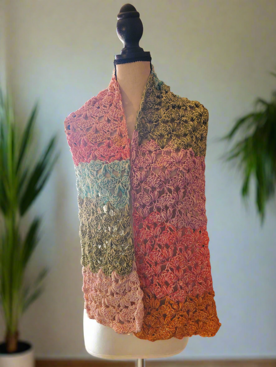 Handmade crochet scarf in soft sunset gradient tones of coral, rose, teal, and golden brown, featuring an intricate lace stitch pattern and lightweight drape, displayed on a dress form in a softly lit room.