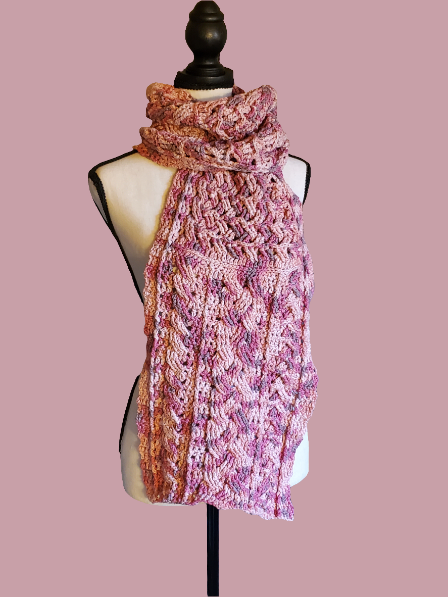 Handmade crochet scarf in soft pink, rose, and peach tones using 100% cotton yarn, featuring a detailed cabled stitch design with a slightly heavier, structured feel, displayed on a dress form.