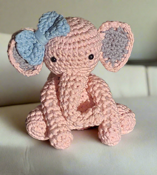 Handmade pink crochet elephant plush with blue bow, soft amigurumi stuffed animal with floppy ears sitting on couch