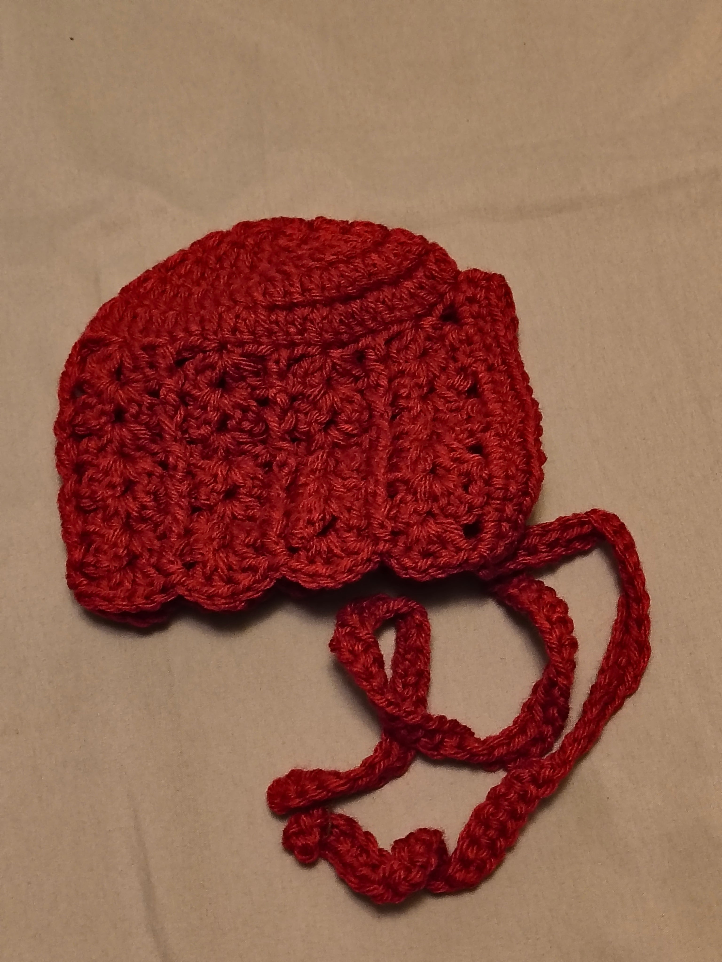 Dark pink crochet baby bonnet with textured stitch pattern, scalloped edge, and tie straps, handmade infant hat laid flat on a neutral background