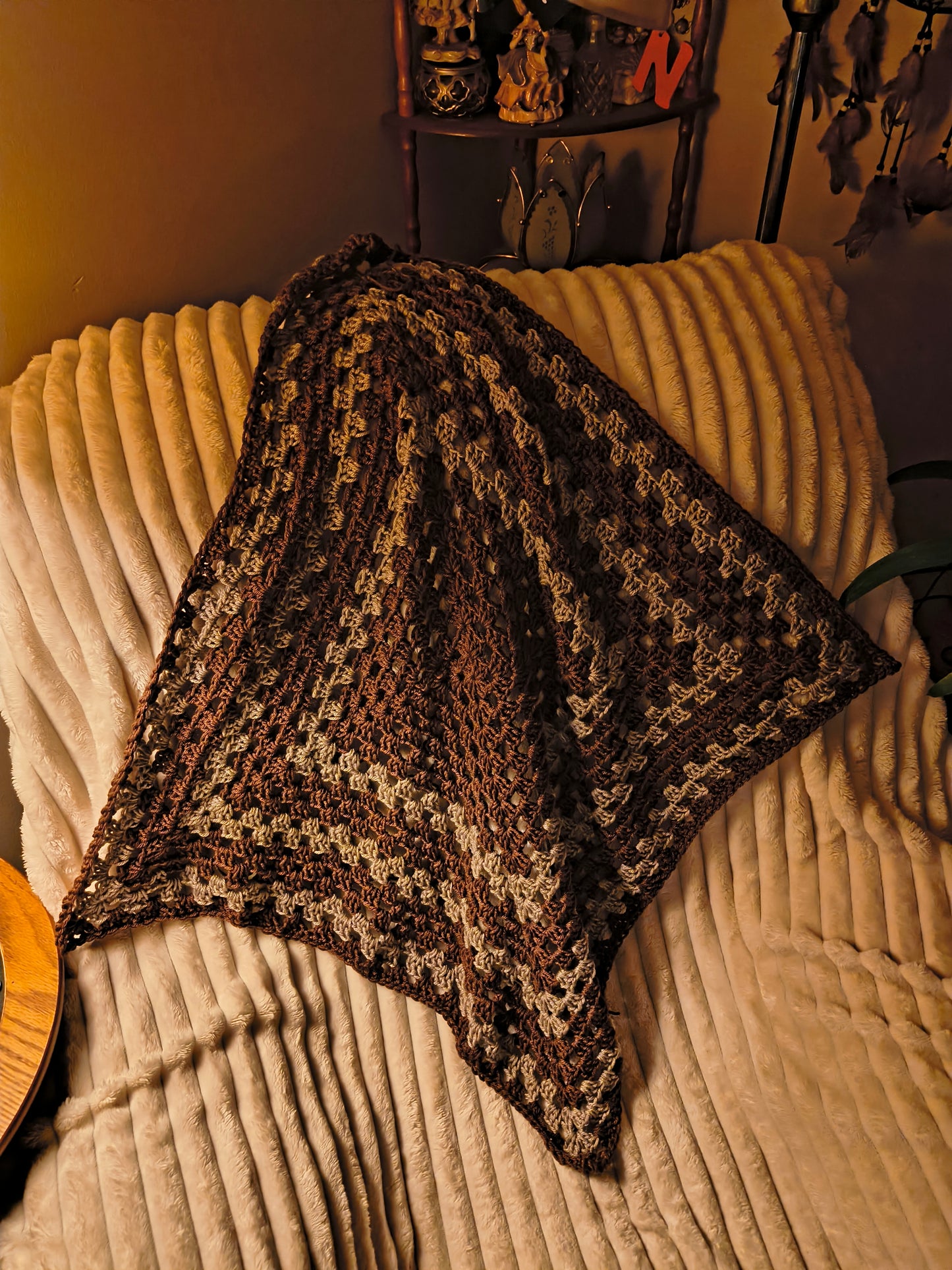 Handmade crochet baby blanket in warm brown and beige tones, featuring a textured granny square pattern, draped over a soft cream ribbed chair in cozy ambient lighting.