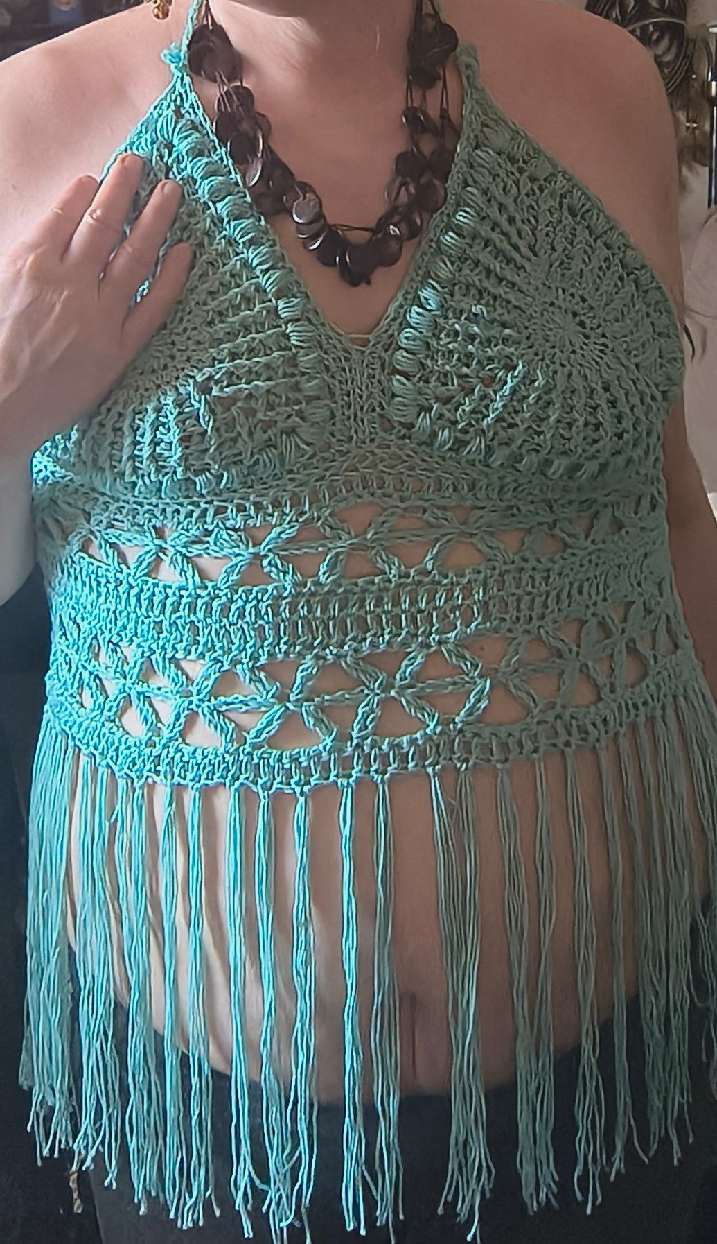 Handmade crochet halter top with textured cups, geometric band detail, and long fringe in teal yarn, worn on body