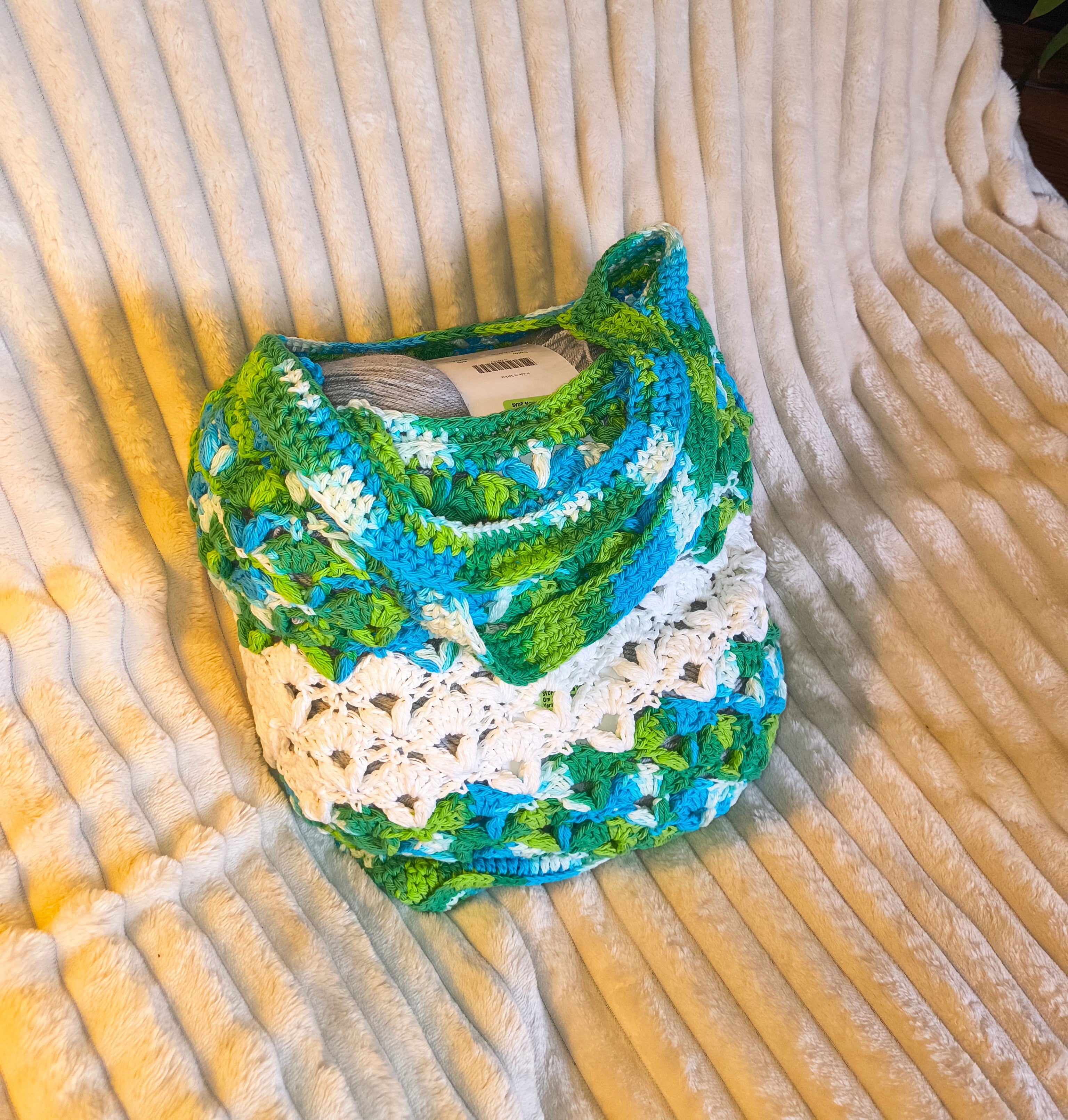 ALT TEXT:
Large handmade crochet market bag in blue, green, and white variegated cotton with camo-inspired pattern and sturdy shoulder straps, displayed on a soft cream blanket