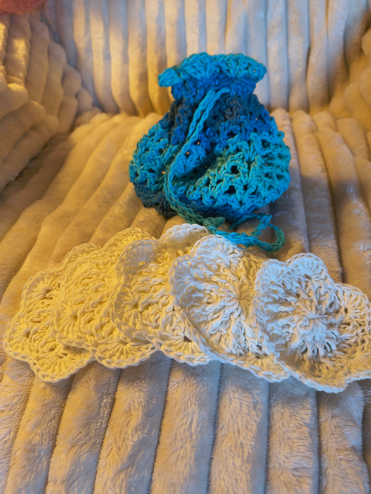 Five cream flower-shaped crochet face scrubbies arranged in front of a blue and green handmade drawstring laundry bag.
