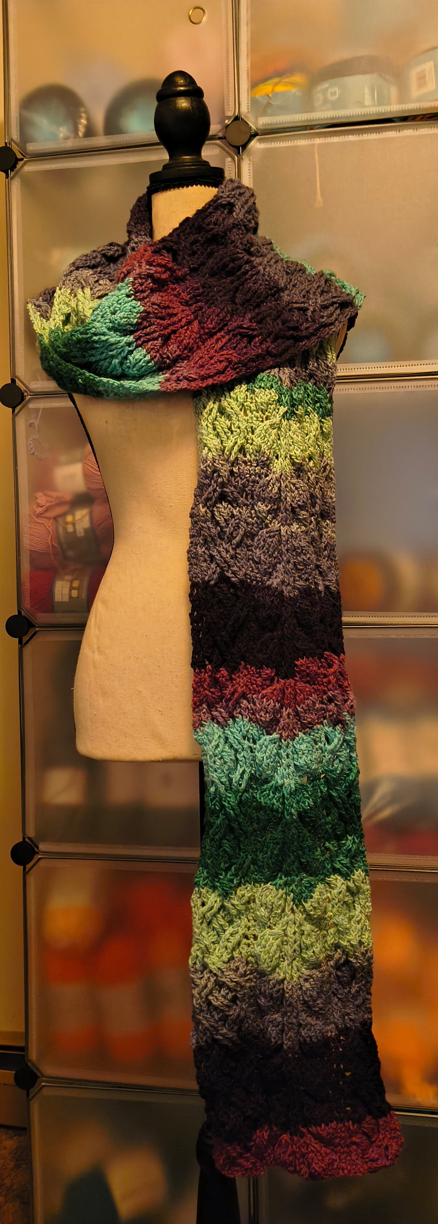 Long handmade crochet super scarf in green, purple, and deep plum tones featuring a textured cabled stitch pattern, draped over a dress form. Extra-long length creates a dramatic, cozy wrap effect with rich color transitions.