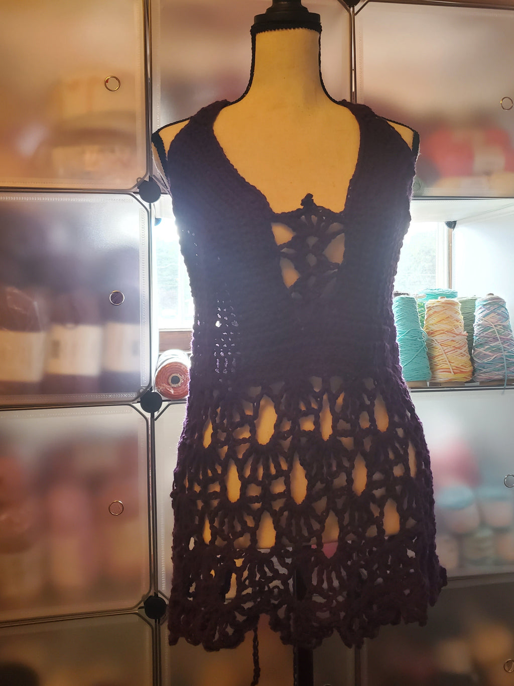 Handmade dark purple crochet top displayed on a dress form, featuring a lace-up front detail, fitted bodice, and an open, flowy lace skirt design, photographed in a cozy craft space with yarn in the background.