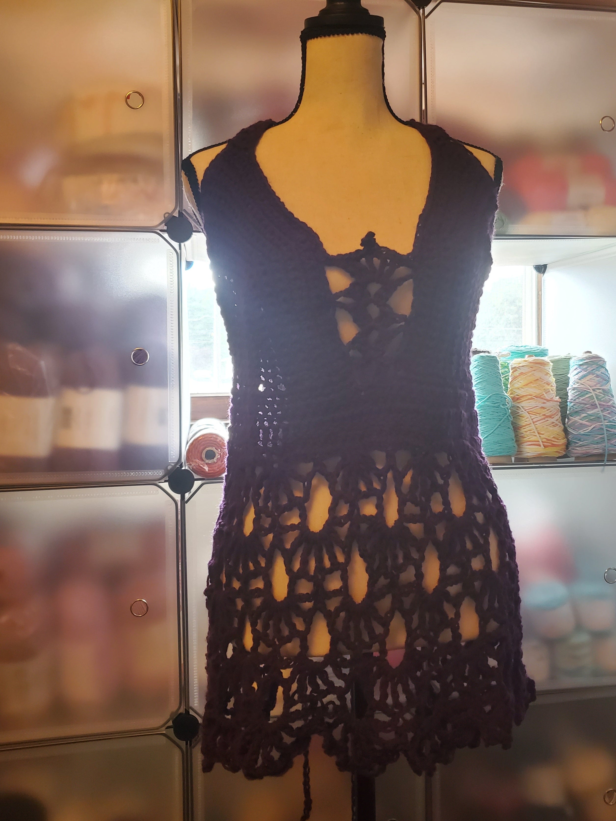 Handmade dark purple crochet top displayed on a dress form, featuring a lace-up front detail, fitted bodice, and an open, flowy lace skirt design, photographed in a cozy craft space with yarn in the background.