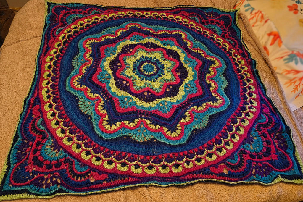 Handmade crochet mandala throw blanket in deep blue, rich teal, vibrant magenta, purple, and bright lime, featuring intricate layered stitch work and bold textured boho design, 55 inch square textile art piece displayed on a bed.