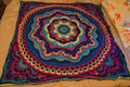 Handmade crochet mandala throw blanket in deep blue, rich teal, vibrant magenta, purple, and bright lime, featuring intricate layered stitch work and bold textured boho design, 55 inch square textile art piece displayed on a bed.
