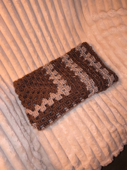 Folded crochet baby blanket in earthy brown and neutral tones, showing detailed stitch texture and soft yarn, displayed on a plush cream surface.