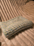 Folded handmade crochet baby blanket in calming blue, green, and white colors displayed on a cozy chair, ideal for stroller or car seat use