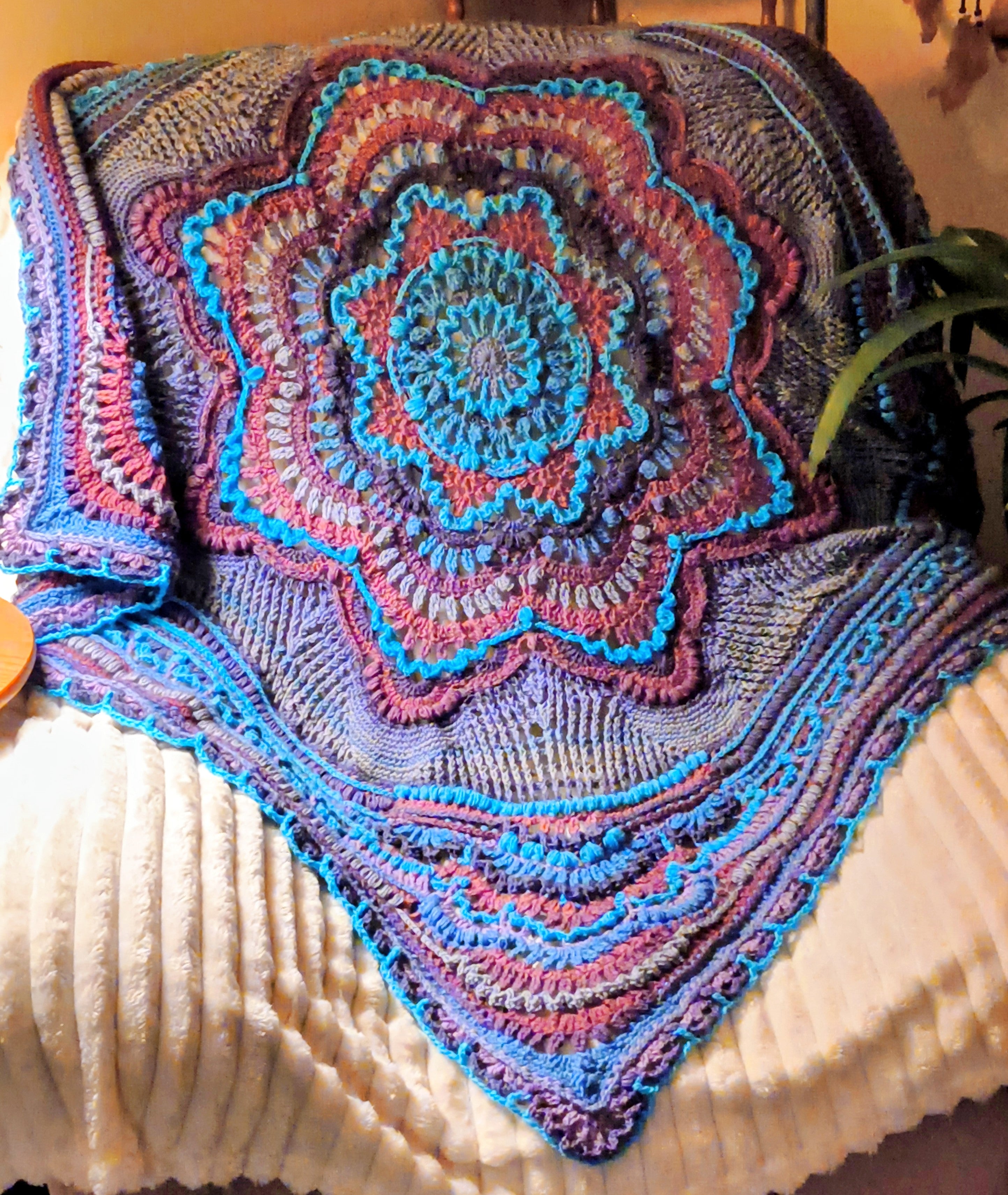 Handcrafted crochet mandala blanket in rich purple, teal, and earthy tones, draped over a cream recliner with a soft textured background, highlighting detailed layered stitches and a symmetrical mandala centerpiece.