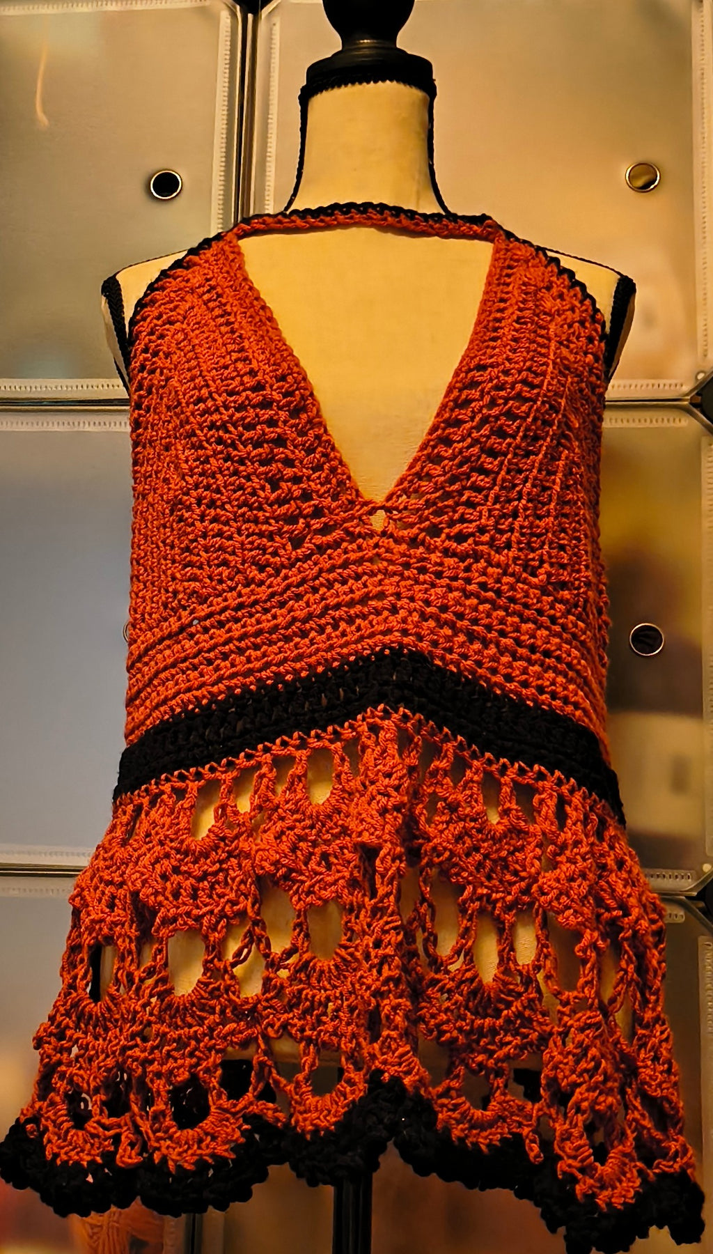 Warm-toned mannequin displaying a handmade crochet halter top in rust orange and black acrylic yarn, featuring textured cups, an open lace lower panel with scalloped edging, and a corset-style lace-up back for an adjustable fit