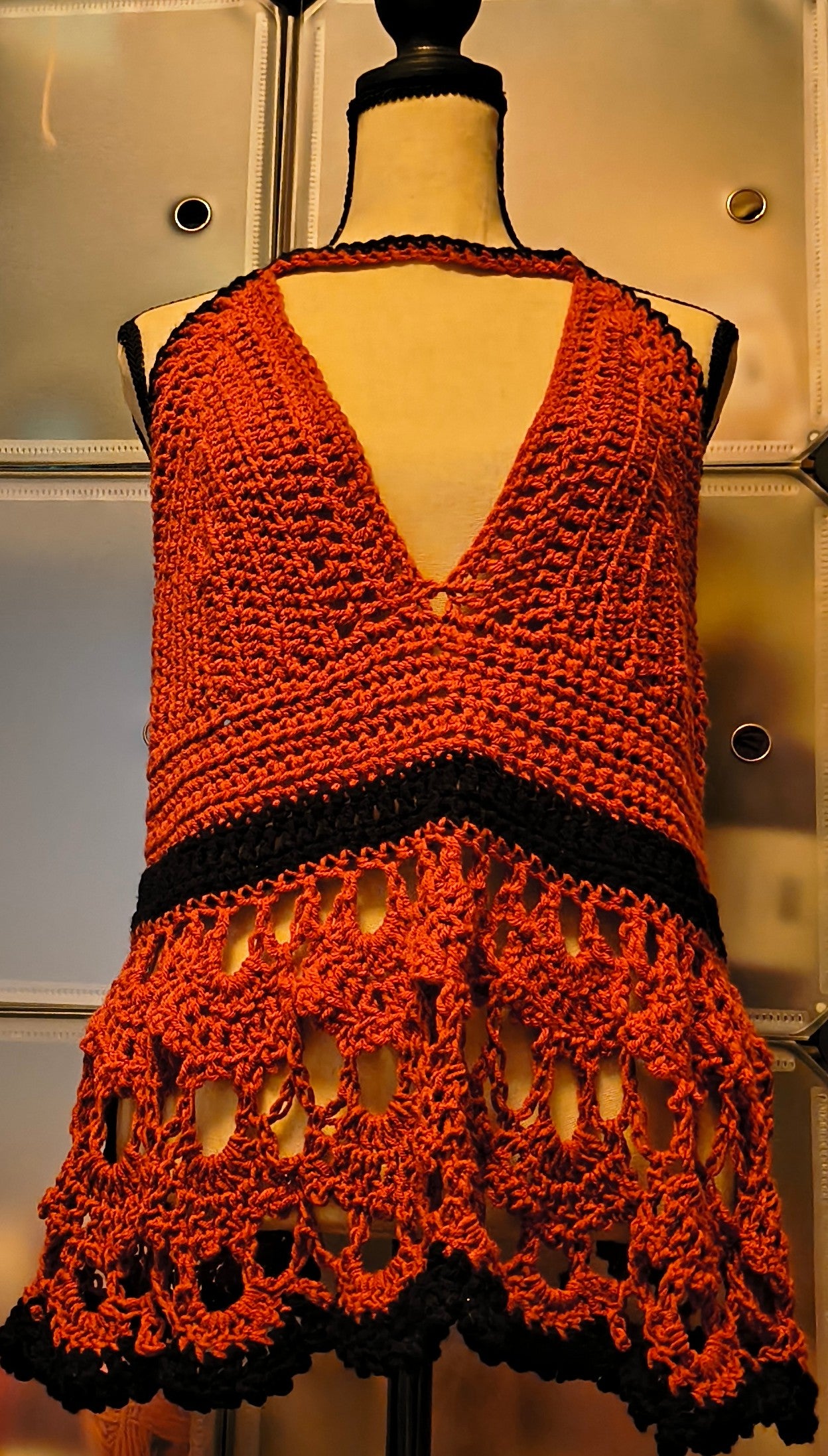 Warm-toned mannequin displaying a handmade crochet halter top in rust orange and black acrylic yarn, featuring textured cups, an open lace lower panel with scalloped edging, and a corset-style lace-up back for an adjustable fit