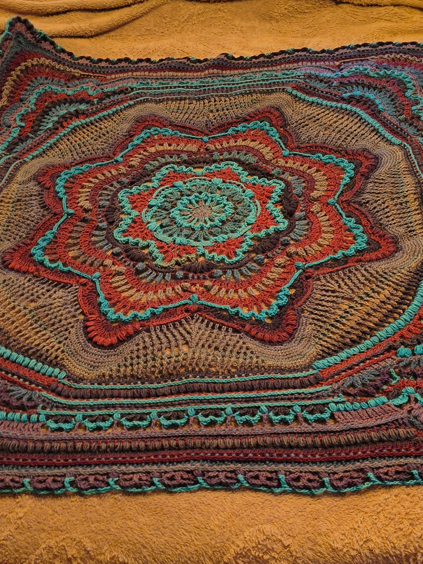 Large handmade crochet mandala blanket in teal, purple, light gray, and dark gray displayed flat on a queen-size bed, featuring intricate layered textures and a bold geometric floral design