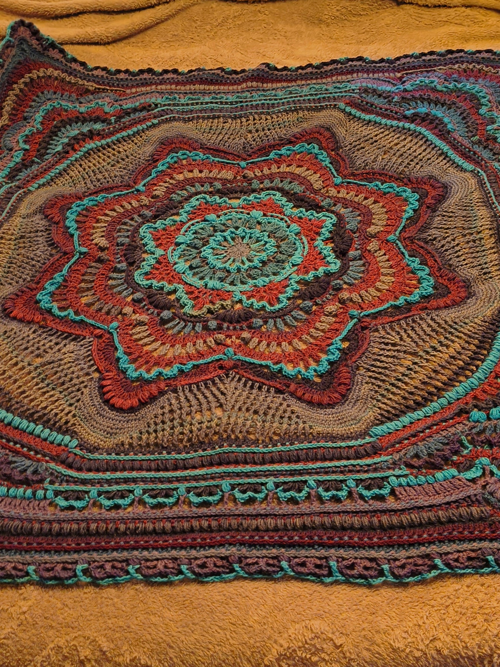 Large handmade crochet mandala blanket in teal, purple, light gray, and dark gray displayed flat on a queen-size bed, featuring intricate layered textures and a bold geometric floral design