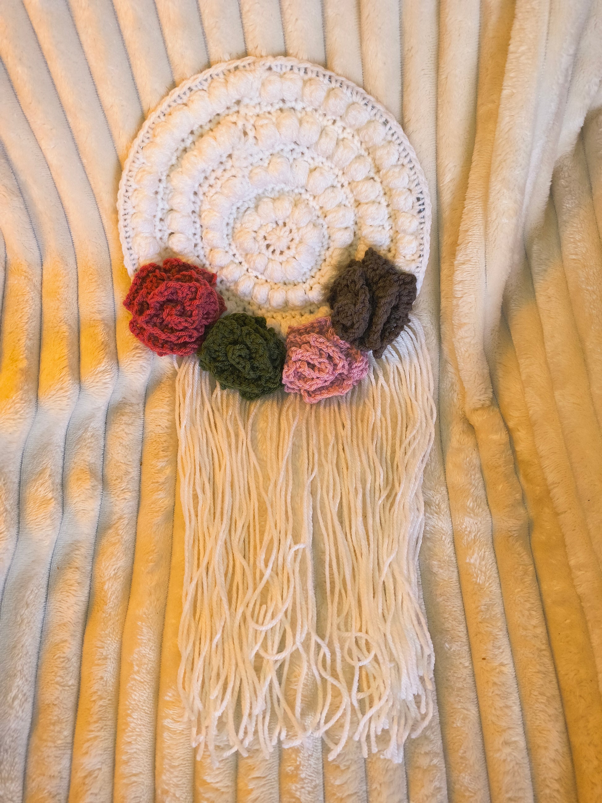Handmade crochet mandala wall hanging in soft white with textured stitching, long fringe, and decorative floral accents in red, green, pink, and brown, displayed on a cozy neutral blanket in a boho style.
