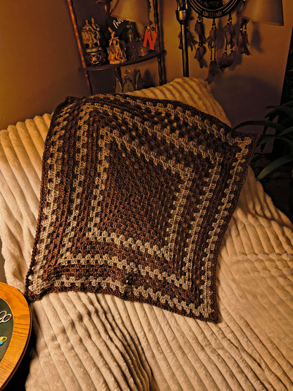 Custom crochet baby blanket in brown and taupe tones with a geometric granny square design, styled on a cream chair with warm, cozy home decor in the background.