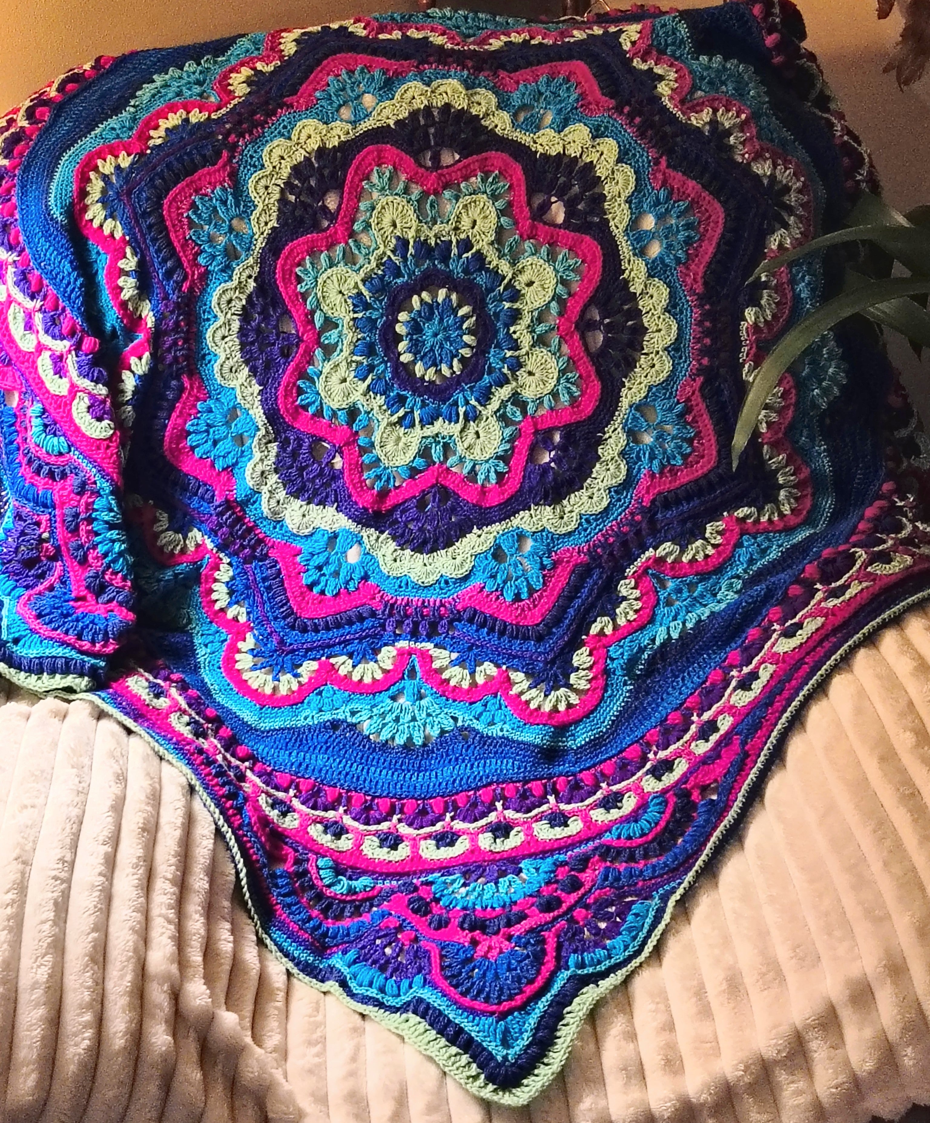 Vibrant crochet mandala blanket draped over a cream recliner, featuring bold pink, teal, blue, and lime green colors with intricate textured stitch patterns and a centered floral mandala design.