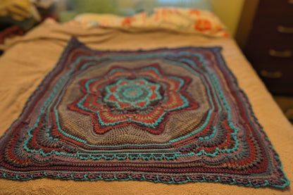 Large handmade crochet mandala blanket in teal, purple, light gray, and dark gray displayed flat on a queen-size bed, featuring intricate layered textures and a bold geometric floral design