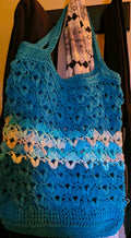 Large handmade crochet market tote in turquoise blue with Evening Sea variegated cotton accents hanging from a coat rack, showing the bag's roomy size, textured stitch pattern, and long shoulder straps.