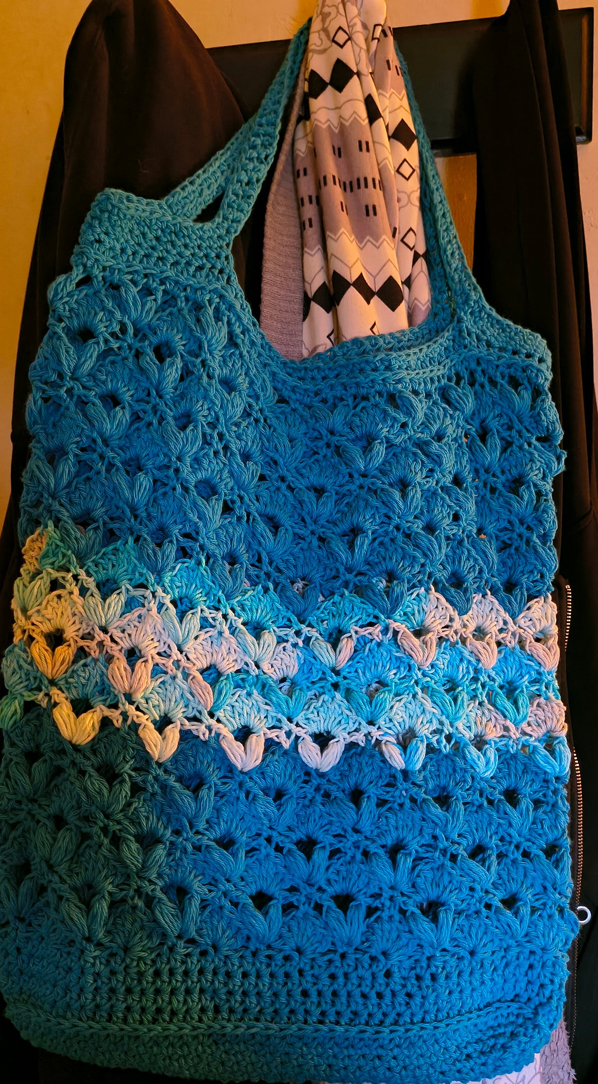 Large handmade crochet market tote in turquoise blue with Evening Sea variegated cotton accents hanging from a coat rack, showing the bag's roomy size, textured stitch pattern, and long shoulder straps.