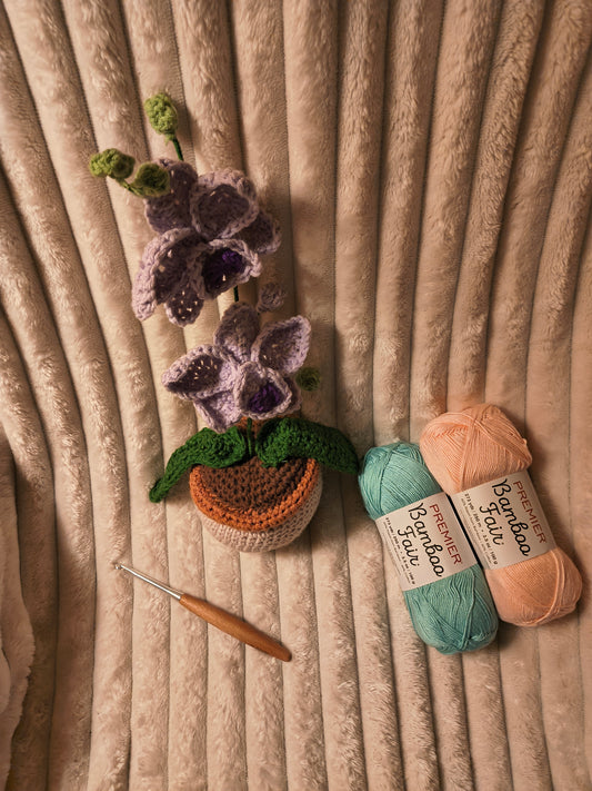 Handmade crochet orchid plant in a small textured pot with purple blossoms and green leaves, displayed on a soft beige blanket alongside a wooden crochet hook and pastel bamboo yarn skeins in mint and peach.